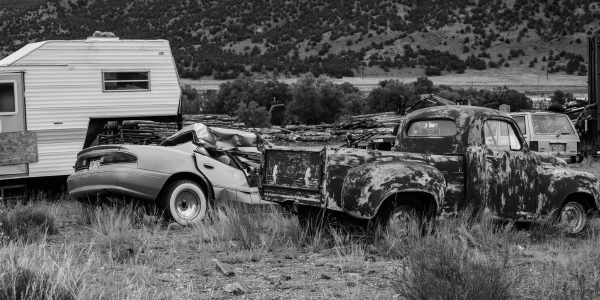 Black and white photo of rusty abandoned vehicles and camper in rural setting.