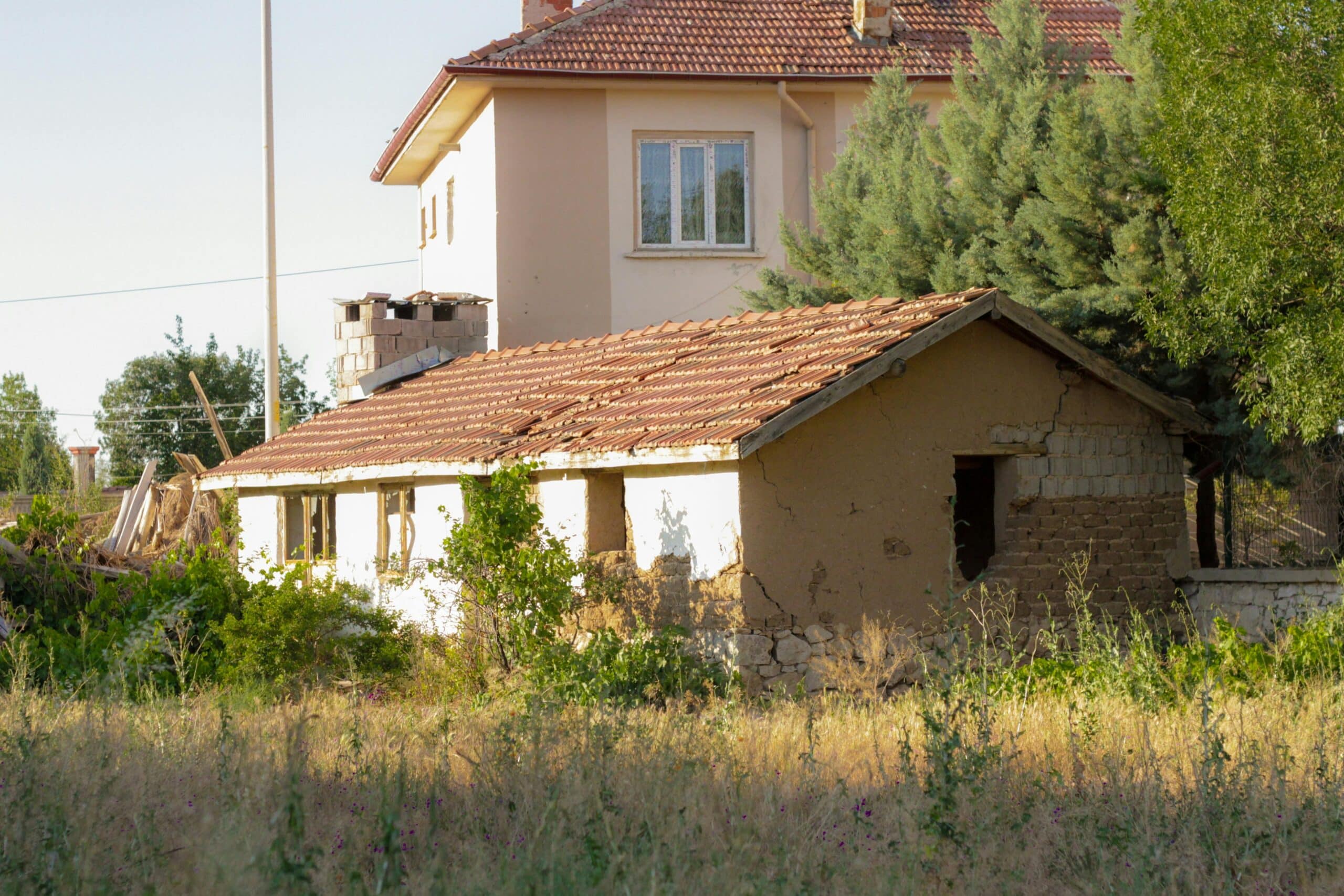 Sunlit abandoned cottage near rural house, depicting rustic charm and decay.